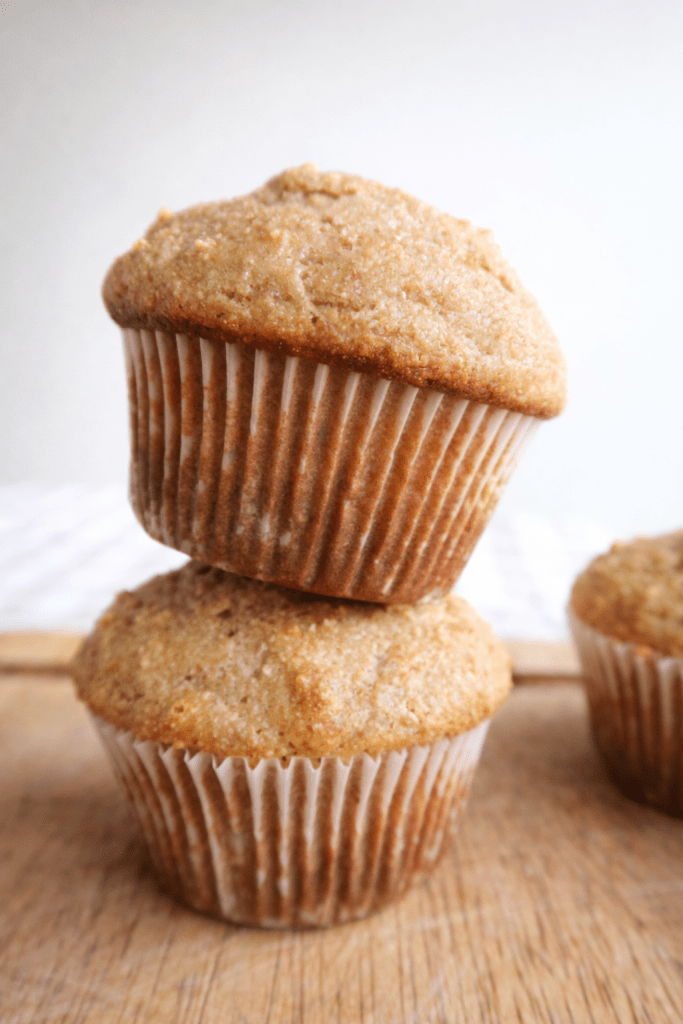 Two freshly baked muffins stacked on a wooden surface, with a third muffin partially visible in the background.