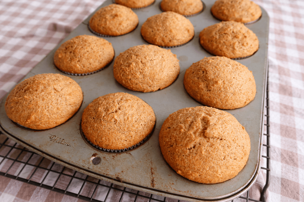 Freshly baked cupcakes cooling in a muffin tin on a checkered cloth.