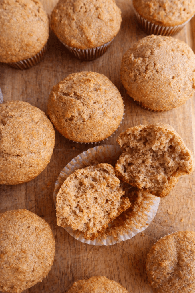 Freshly baked whole grain muffins on a wooden surface, with one muffin cut in half to reveal its soft, textured interior.