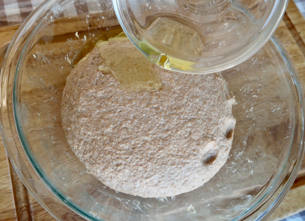 A glass bowl containing a round dough with a small amount of oil being poured on top.