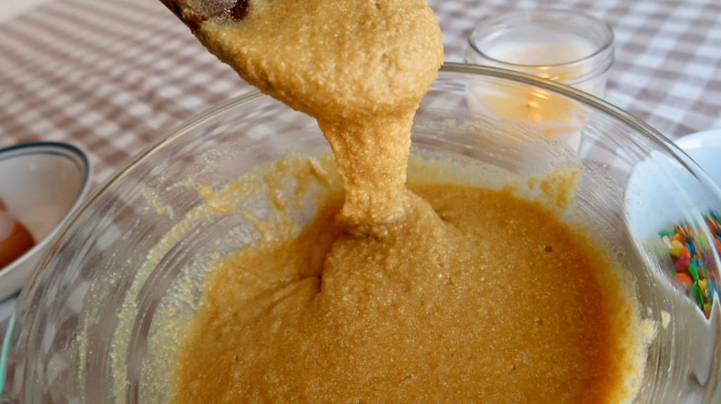 Close-up of a wooden spatula scooping thick, golden batter from a glass mixing bowl, with eggs and colorful sprinkles visible in the background.