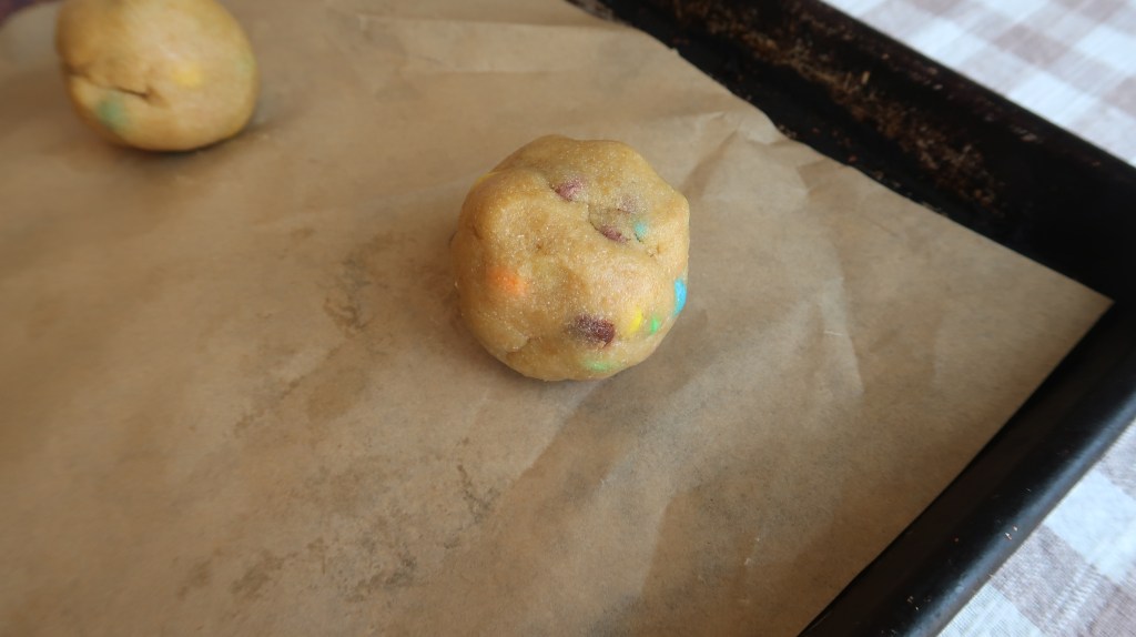 A close-up view of cookie dough balls on parchment paper, with colorful chocolate chips visible in the dough.