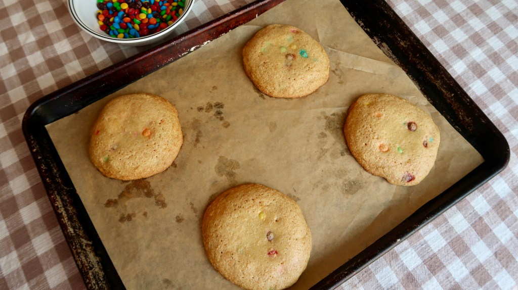 Freshly baked cookies with colorful chocolate pieces on a baking tray lined with parchment paper, accompanied by a bowl of candy on a checkered tablecloth.