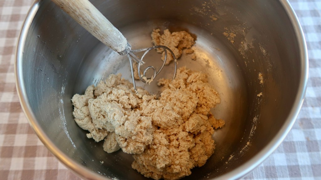 A metal mixing bowl containing a lump of dough and a metal mixing attachment with a wooden handle, placed on a checkered tablecloth.