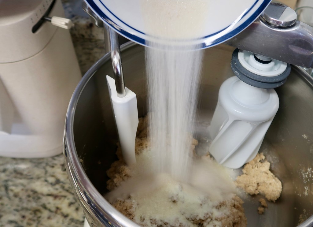 Sugar being poured into a mixing bowl with cookie dough and a mixer in the background.