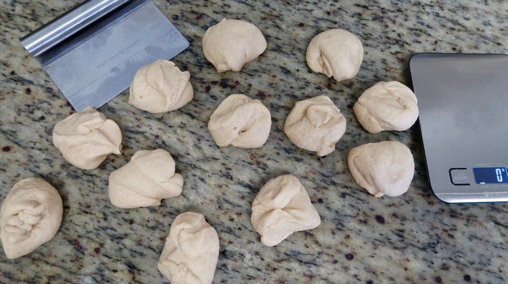 Several small dough balls arranged on a granite countertop next to a kitchen scale and a dough scraper.
