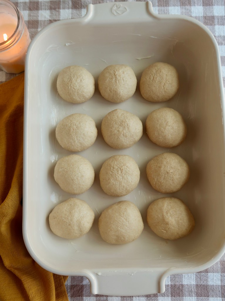 A baking dish with twelve rounded dough balls, arranged evenly in a grid pattern, on a checkered tablecloth.