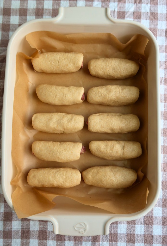 A baking dish lined with parchment paper containing freshly rolled dough filled with hot dogs, arranged neatly in two rows.