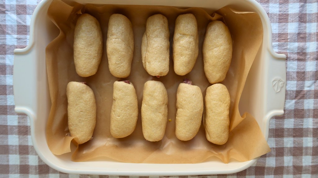 A baking dish containing ten small, oval-shaped dough pieces arranged in two rows, placed on brown parchment paper.