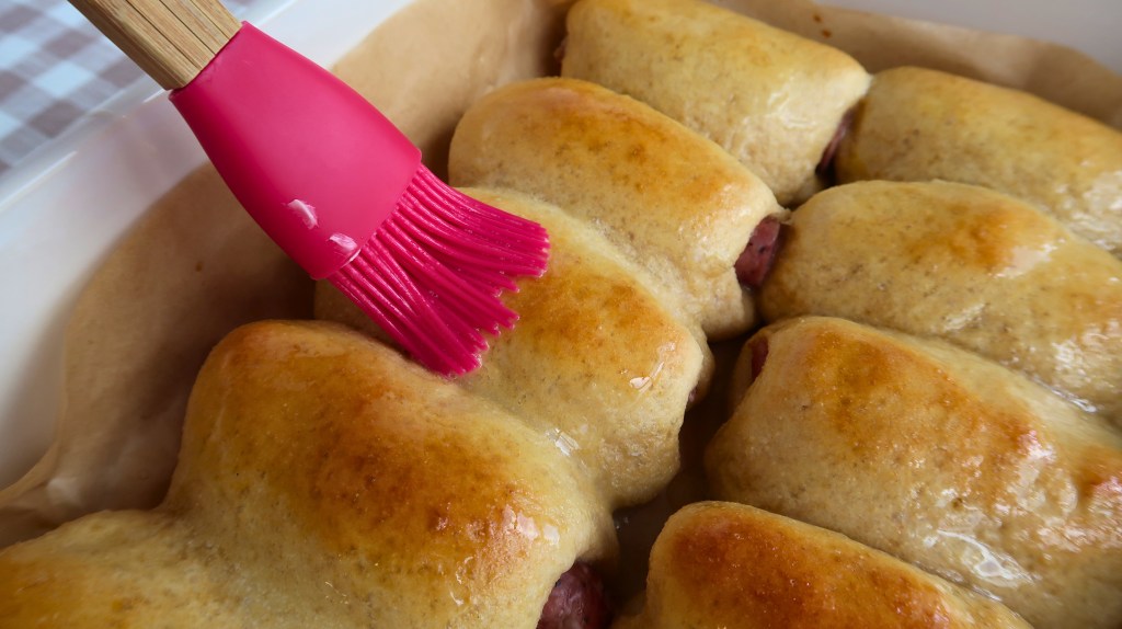 A close-up of freshly baked rolls being brushed with egg wash using a pink silicone brush.