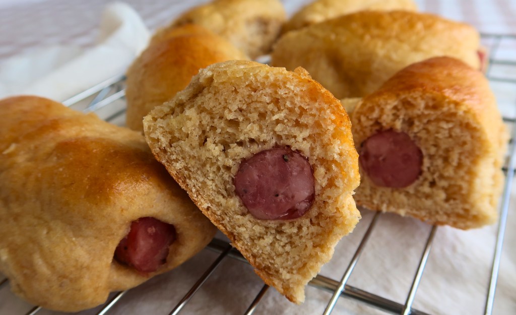 Close-up of freshly baked bread rolls with sliced hot dogs inside, arranged on a wire rack.