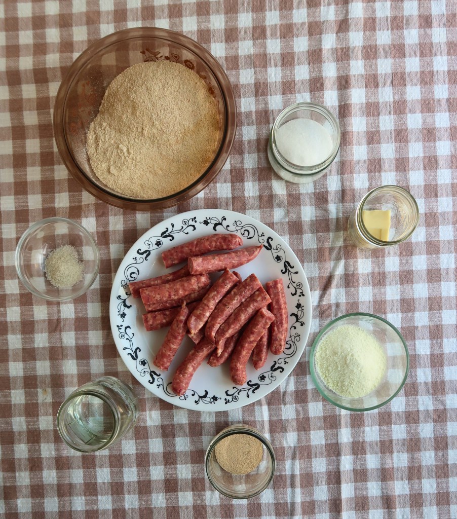 An assortment of cooking ingredients arranged on a checkered tablecloth, including a bowl of flour, a plate of sausage links, and various jars containing sugar, butter, and spices.