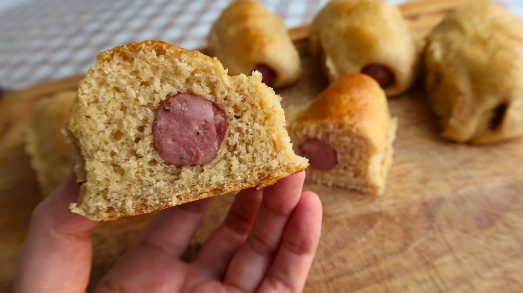 A hand holding a sliced piece of bread with a hot dog inside, surrounded by whole baked rolls on a wooden cutting board.