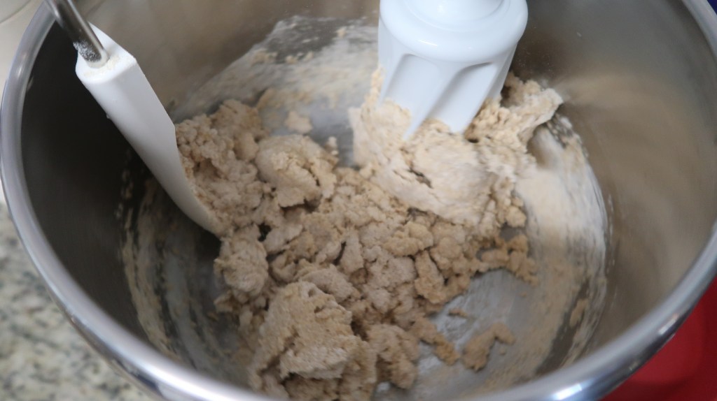 A metal mixing bowl containing dough being mixed with a paddle attachment.