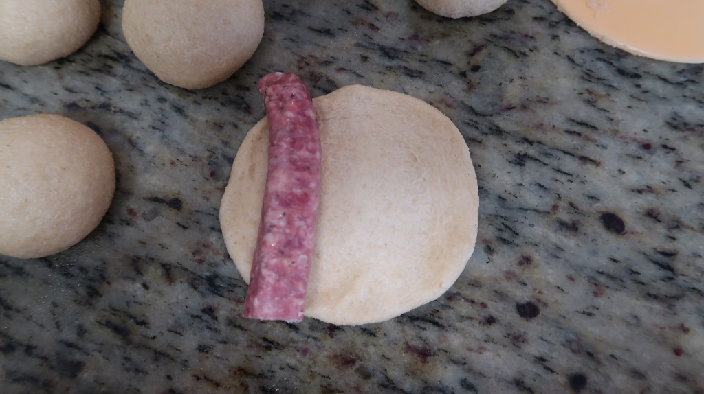 A piece of uncooked dough with a sausage placed on top, surrounded by small dough balls on a granite countertop.