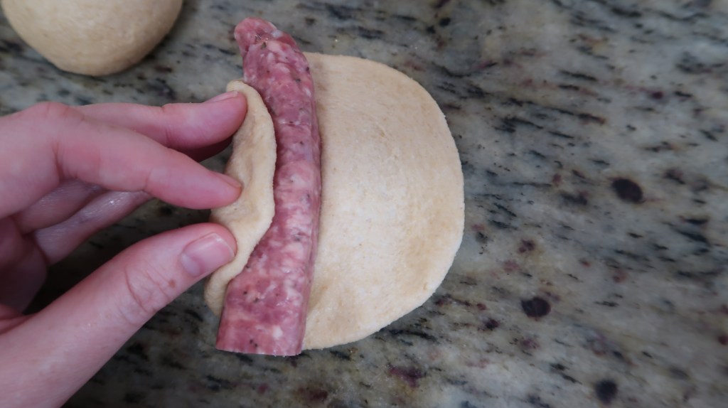 A hand folding dough around a sausage on a textured countertop.