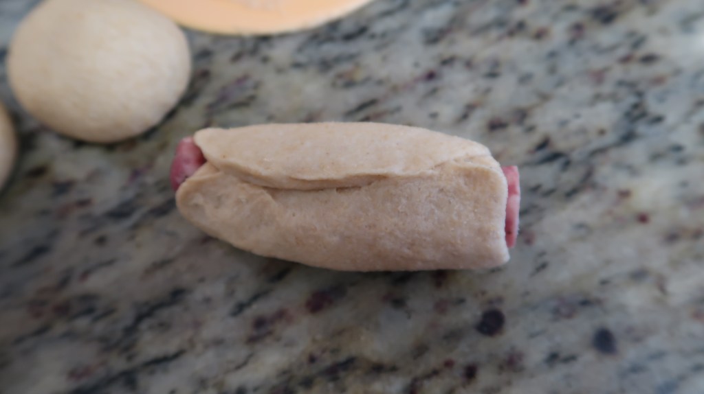 A uncooked hot dog wrapped in dough, resting on a granite countertop with round dough balls nearby.